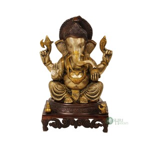 May include: A gold and silver Ganesha statue sitting on a wooden base with intricate carvings. The statue is holding a small axe and a lotus flower in its hands. The base has a small elephant figurine on one side and the text "Shree Handicrafts" on the other.