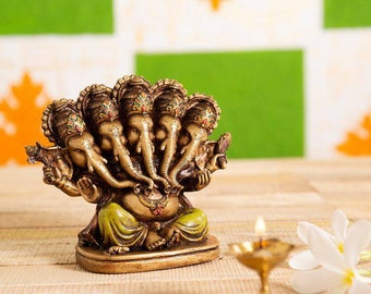 May include: A gold-colored statue of Ganesha with five heads and multiple arms. The statue is sitting on a base and has a green and gold colored garment. The statue is in front of a green and white background.