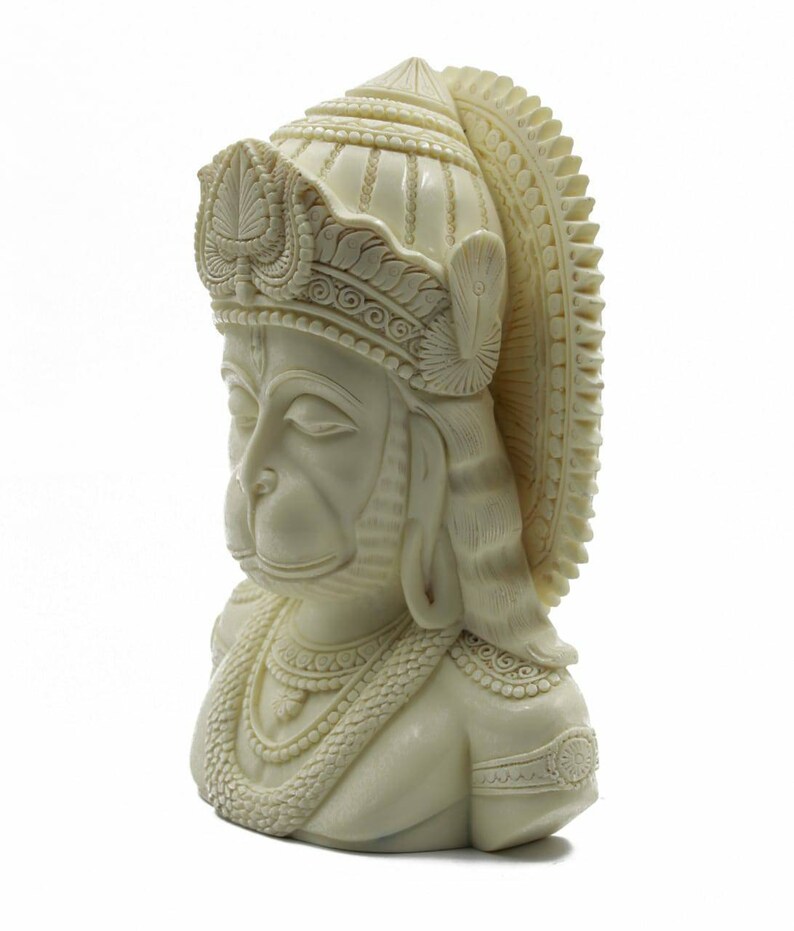 May include: A white, detailed bust sculpture of the Hindu deity Hanuman. The sculpture depicts Hanuman with a crown and a necklace, and his eyes are closed.