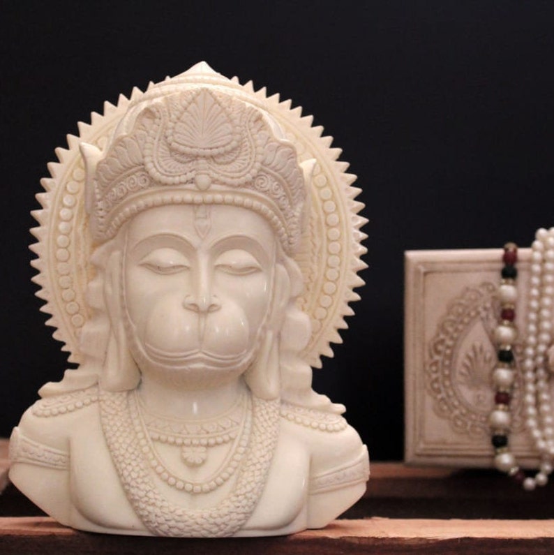 May include: White ivory bust sculpture of the Hindu deity Hanuman, with a crown and a halo. The sculpture is detailed with intricate carvings and is a traditional religious art piece.