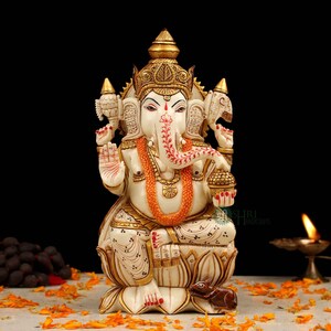May include: White Ganesha statue with gold accents, sitting on a lotus base with a mouse at its feet. The statue is decorated with intricate details and has a red and white floral pattern on its trunk.