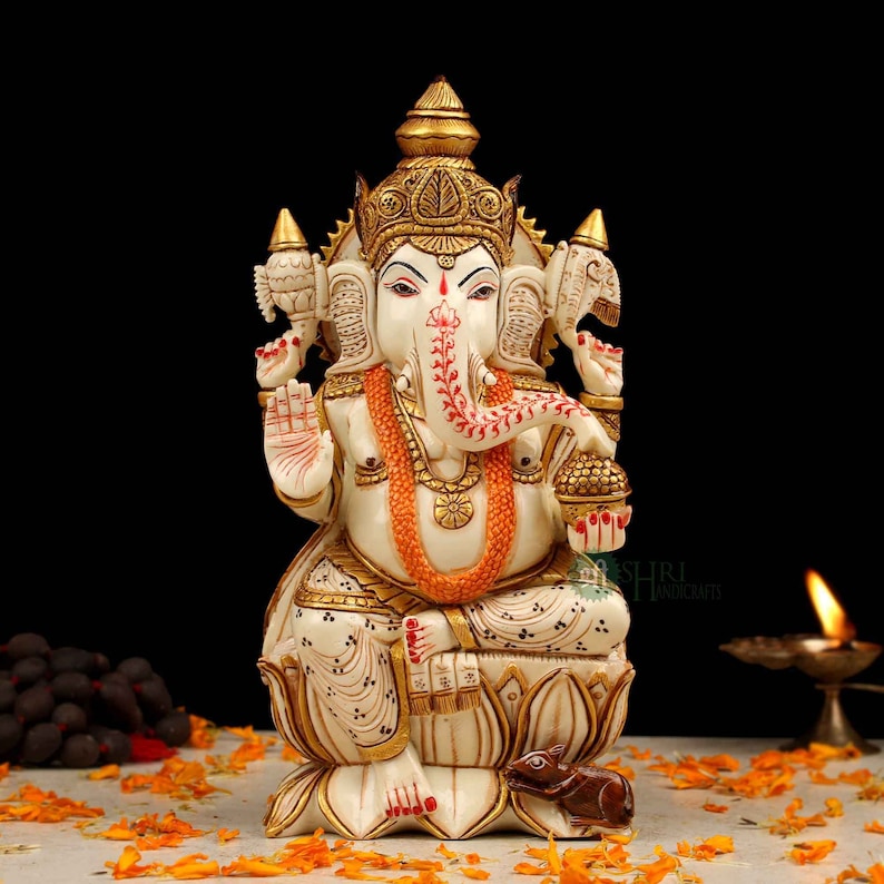 May include: White Ganesha statue with gold accents, sitting on a lotus base with a mouse at its feet. The statue is decorated with intricate details and has a red and white floral pattern on its trunk.