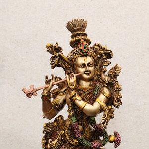 May include: A gold-colored statue of Krishna playing a flute. The statue is intricately detailed with ornate jewelry and a crown. The background is a plain white wall.