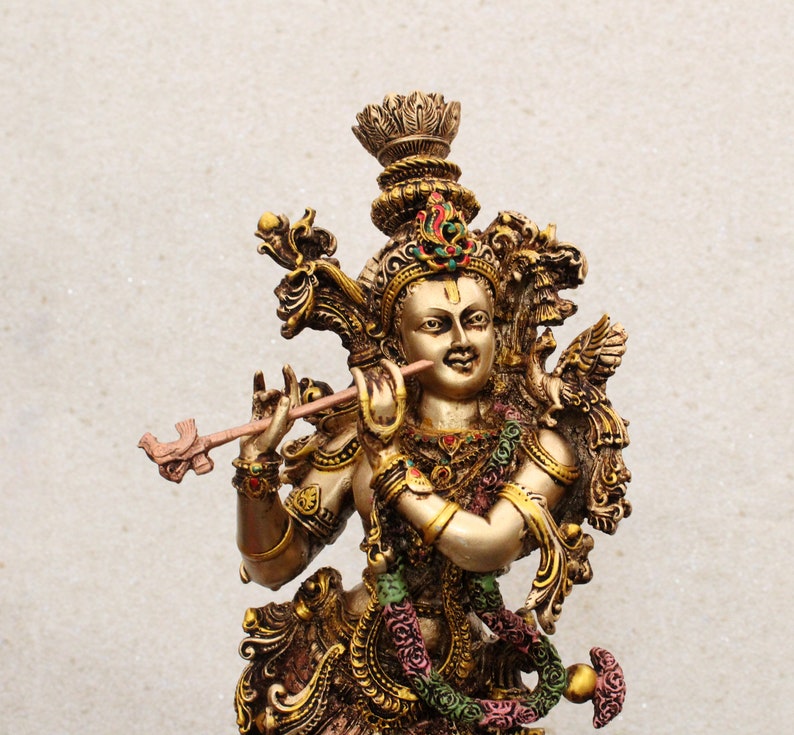 May include: A gold-colored statue of Krishna playing a flute. The statue is intricately detailed with ornate jewelry and a crown. The background is a plain white wall.