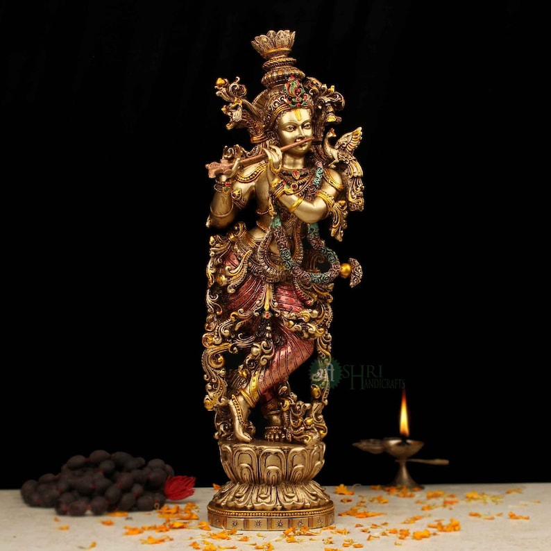 May include: A gold-toned statue of Krishna playing a flute, standing on a lotus base. The statue is intricately detailed with colorful accents and floral patterns.