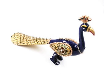 Puede incluir: Una figurita de pavo real azul con una cola incrustada de oro y perlas. El pavo real está decorado con joyas de colores y tiene una corona dorada.