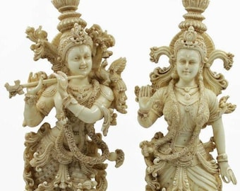 May include: A pair of intricately carved ivory-colored statues depicting Hindu deities Radha and Krishna. Krishna is playing a flute and Radha is holding a flower in her hand. The statues are adorned with elaborate jewelry and clothing.
