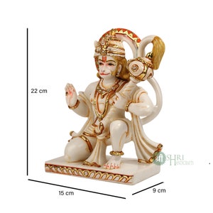 Może przedstawiać: Biała marmurowa statua hinduskiego b&oacute;stwa Hanuman, siedzącego z uniesioną prawą ręką w geście błogosławieństwa. Trzyma maczugę w lewej ręce. Statua jest ozdobiona złotą farbą i ma czerwoną i pomarańczową aureolę wok&oacute;ł głowy. Statua ma 22 cm wysokości, 15 cm szerokości i 9 cm głębokości.