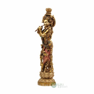 May include: A gold-toned statue of Krishna playing a flute, standing on a decorative base. The statue is adorned with intricate details and colorful accents.