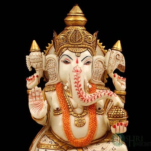 May include: White marble statue of Ganesha, the Hindu god of wisdom and prosperity. The statue is intricately carved with gold accents and features a red and white floral design on the trunk. Ganesha is depicted with four arms, holding a broken tusk, a sweetmeat, a lotus flower, and an axe.
