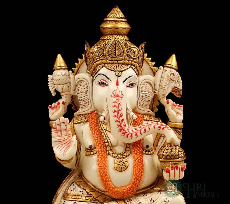 May include: White marble statue of Ganesha, the Hindu god of wisdom and prosperity. The statue is intricately carved with gold accents and features a red and white floral design on the trunk. Ganesha is depicted with four arms, holding a broken tusk, a sweetmeat, a lotus flower, and an axe.