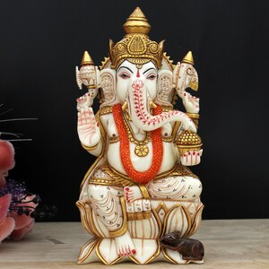May include: White marble statue of Ganesha, the Hindu god of wisdom and prosperity, sitting on a lotus flower. The statue is decorated with gold paint and red accents. A small mouse is at the base of the statue.