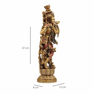 May include: A gold-toned statue of a Hindu deity playing a flute, standing on a lotus-shaped base. The statue is 37 cm tall, 12 cm wide, and 8 cm deep.