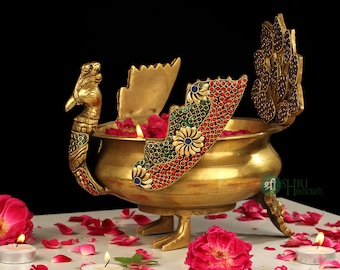 Puede incluir: Un quemador de incienso de color dorado con forma de pavo real con detalles intrincados y piedras preciosas de colores. El quemador está decorado con piedras rojas, verdes y azules y tiene un cuenco redondo para quemar incienso. El quemador está sentado sobre una superficie blanca con pétalos de rosa esparcidos a su alrededor.