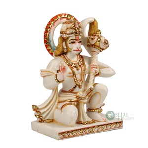 Może przedstawiać: Biała marmurowa statua hinduskiego b&oacute;stwa Hanuman, siedzącego z uniesioną prawą ręką w geście błogosławieństwa. Trzyma maczugę w lewej ręce. Statua jest ozdobiona złotą farbą i ma czerwoną i złotą aureolę za głową. Statua stoi na białej podstawie ze złotą obw&oacute;dką.
