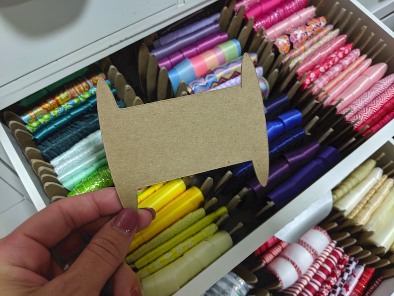 Puede incluir: Un carrete de cart&oacute;n para cinta, con una variedad de cintas de colores en el fondo. Las cintas est&aacute;n organizadas en un caj&oacute;n e incluyen colores como rosa, morado, azul, verde, amarillo y blanco.