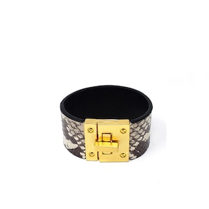 May include: A wide, brown leather cuff bracelet with a gold-tone clasp. The bracelet has a snakeskin pattern.