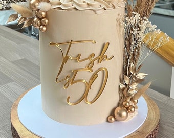 Pode incluir: Um bolo bege decorado com detalhes dourados e flores secas. O bolo está em um suporte de bolo de madeira e diz "Fresh 50" no lado.