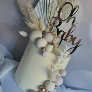 Puede incluir: Un pastel blanco decorado con esferas blancas y doradas, hierba de la pampa seca y un adorno de pastel dorado 'Oh Baby'.