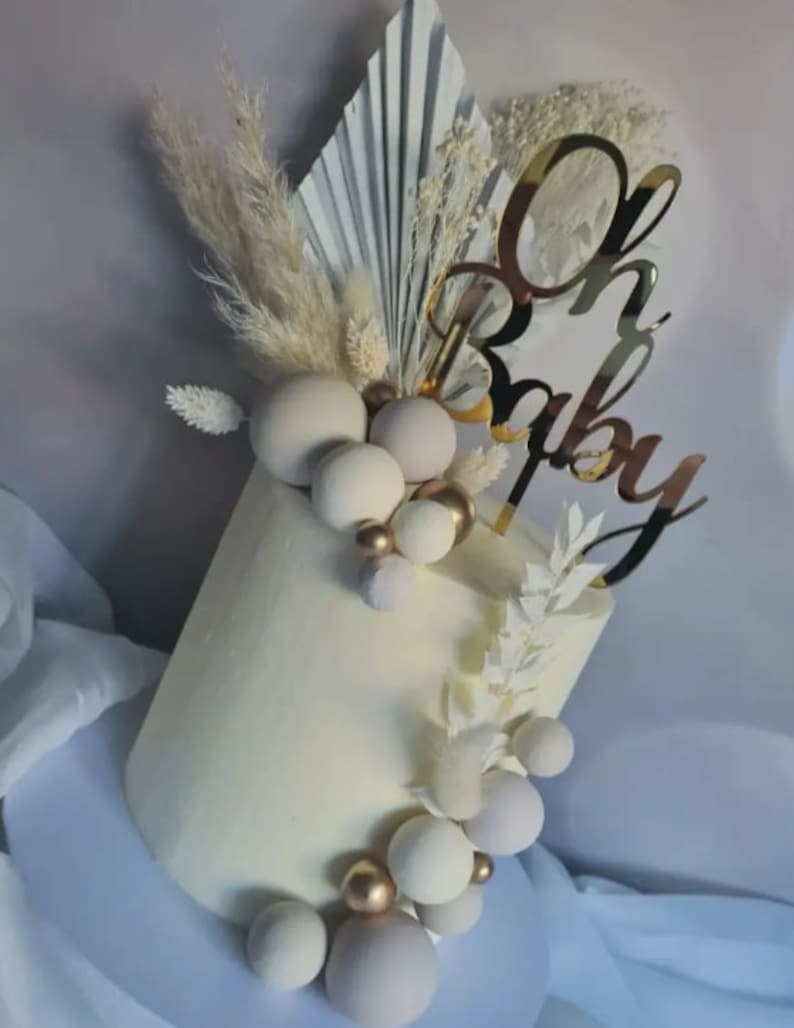 Puede incluir: Un pastel blanco decorado con esferas blancas y doradas, hierba de la pampa seca y un adorno de pastel dorado 'Oh Baby'.