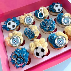 Puede incluir: Una caja rosa llena de cupcakes decorados. Algunos tienen glaseado azul con adornos de bal&oacute;n de f&uacute;tbol. Otros tienen glaseado blanco con una insignia de f&uacute;tbol o el n&uacute;mero 8. Decoraciones plateadas.