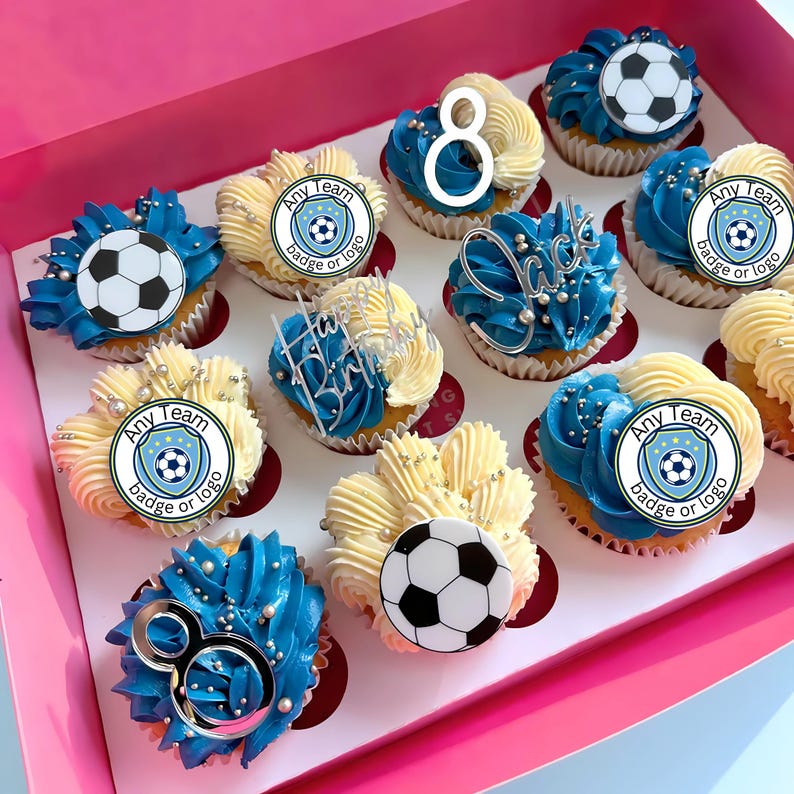 Puede incluir: Una caja rosa llena de cupcakes decorados. Algunos tienen glaseado azul con adornos de bal&oacute;n de f&uacute;tbol. Otros tienen glaseado blanco con una insignia de f&uacute;tbol o el n&uacute;mero 8. Decoraciones plateadas.