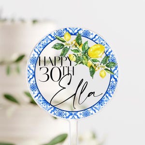 May include: A clear, round cake topper with a blue and white patterned border. The topper features the text "HAPPY 30TH Ella" with a watercolor lemon and leaf design. The topper is designed to be inserted into a cake.