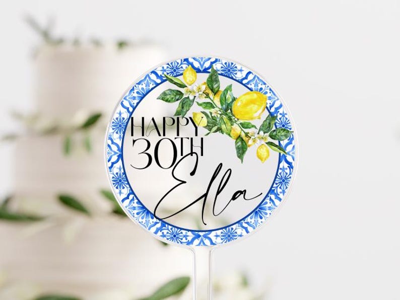May include: A clear, round cake topper with a blue and white patterned border. The topper features the text "HAPPY 30TH Ella" with a watercolor lemon and leaf design. The topper is designed to be inserted into a cake.