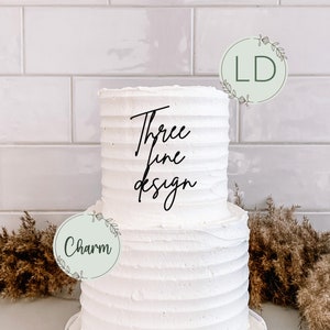 May include: A two-tier white cake with buttercream frosting and a simple, elegant design. The cake is decorated with two round, green paper circles with white text. The top circle says "LD" and the bottom circle says "Charm". The cake is also decorated with black cursive text that says "Three Fine Design".