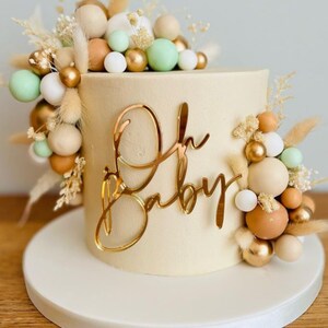 Puede incluir: Un pastel blanco con un adorno dorado de "Oh Baby" y una decoraci&oacute;n de bolas beige, marr&oacute;n, dorado y verde y flores secas.