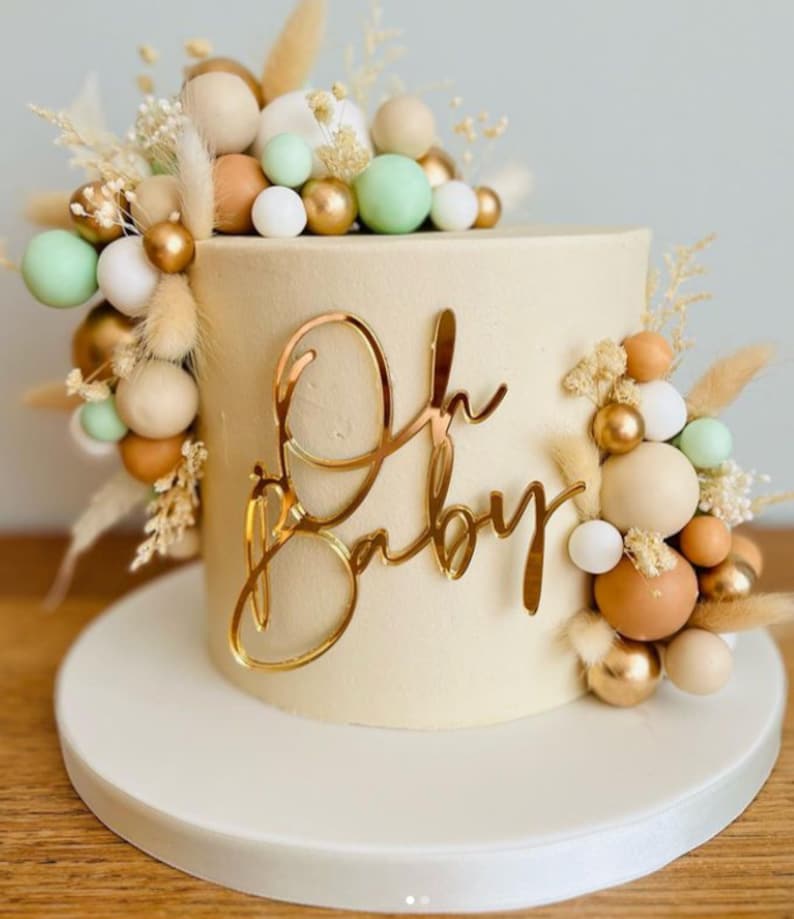 Puede incluir: Un pastel blanco con un adorno dorado de "Oh Baby" y una decoraci&oacute;n de bolas beige, marr&oacute;n, dorado y verde y flores secas.