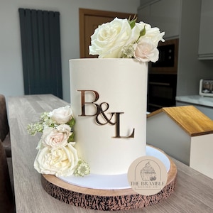 May include: A white wedding cake with a simple design, decorated with white roses and greenery. The cake is topped with gold letters spelling "B&L" and a small round sticker with the text "THE BRAMBLE BAKING CO."