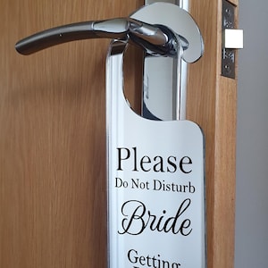 May include: A white door hanger with the text "Please Do Not Disturb Bride Getting Ready" in black script is hanging on a door handle. The door handle is chrome and the door is made of wood.