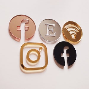 May include: Five different social media icons made of acrylic. The icons are in the shape of a Facebook 'f', an Instagram camera, a Twitter bird, a WiFi symbol, and a letter 'E'. The icons are all different colours, including gold, silver, black, and rose gold.