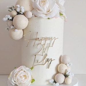 May include: A tall, cylindrical cake with smooth off-white frosting and a speckled finish. It is decorated with clusters of beige spheres, small white flowers, and large white roses. A gold-coloured acrylic topper reads Happy 21st Lily.