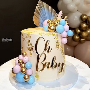 Puede incluir: Un pastel blanco decorado con pan de oro y una decoraci&oacute;n dorada de 'Oh Baby'. El pastel est&aacute; cubierto con un grupo de globos rosas y azules.