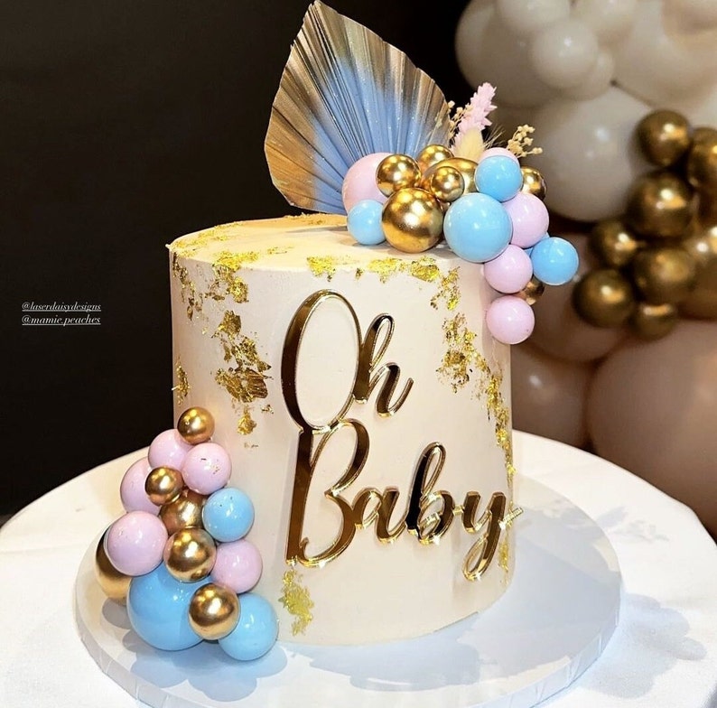 Puede incluir: Un pastel blanco decorado con pan de oro y una decoraci&oacute;n dorada de 'Oh Baby'. El pastel est&aacute; cubierto con un grupo de globos rosas y azules.