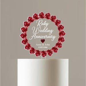May include: A circular cake topper featuring a border of red faceted gems. The centre is clear with white text reading Ruby Wedding Anniversary, a small red heart, and the names Anne & John with the date 15/07/1986 printed below the heart.