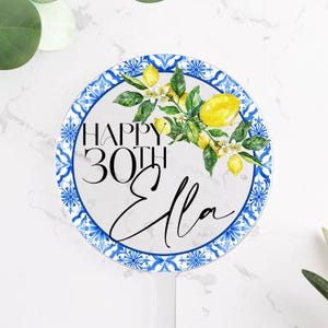 May include: A round, white cake topper with a blue and white patterned border. The topper features the text "HAPPY 30TH Ella" with a watercolor lemon and floral design. The topper is on a white surface with greenery.