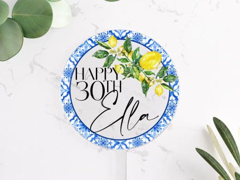 May include: A round, white cake topper with a blue and white patterned border. The topper features the text "HAPPY 30TH Ella" with a watercolor lemon and floral design. The topper is on a white surface with greenery.