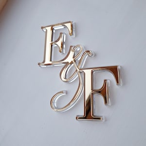 May include: A decorative sign featuring the letters E and F in a serif font, connected by an ampersand symbol. The letters are constructed from a clear acrylic base with a reflective, metallic gold-toned finish on the front surface.