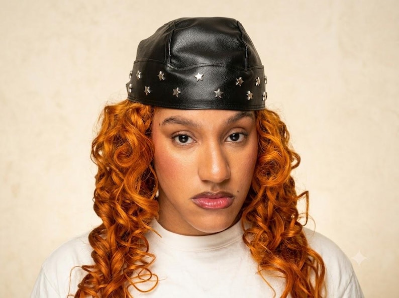 May include: A black faux leather skull cap featuring a band of small, silver-toned metal star-shaped studs encircling the base. The cap has a smooth, structured finish and sits firmly on the head, showcasing a clean, minimalist design.