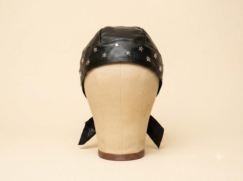 May include: A black head covering featuring a textured surface and a row of small, silver-toned metal star-shaped studs arranged along the front edge. The head covering includes two fabric ties extending from the sides for securing the item.