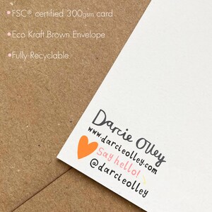 May include: A white card with a pink heart and the text "Darcie Olley" in black. The text "www.darcieolley.com" is below the name in black. The text "Say hello!" is below the website address in black. The text "@darcieolley" is below the greeting in black. The card is on a brown surface.