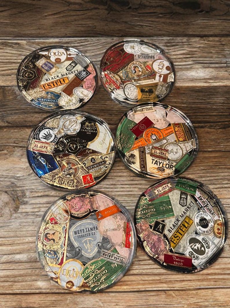 Puede incluir: Seis posavasos redondos con tapas de resina transparente, que muestran un collage de etiquetas de cigarros antiguos. Las etiquetas presentan varios colores, fuentes y dise&ntilde;os, incluyendo textos como "OLIVA", "BLACK MARKET" y "ESTELI". Los posavasos est&aacute;n colocados sobre una superficie de madera.