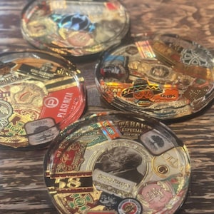 Puede incluir: Cuatro posavasos de resina transparente con dise&ntilde;os de etiquetas de cigarros. Cada posavasos presenta una etiqueta diferente con detalles dorados, rojos y marrones. Los posavasos est&aacute;n dispuestos sobre una superficie de madera.
