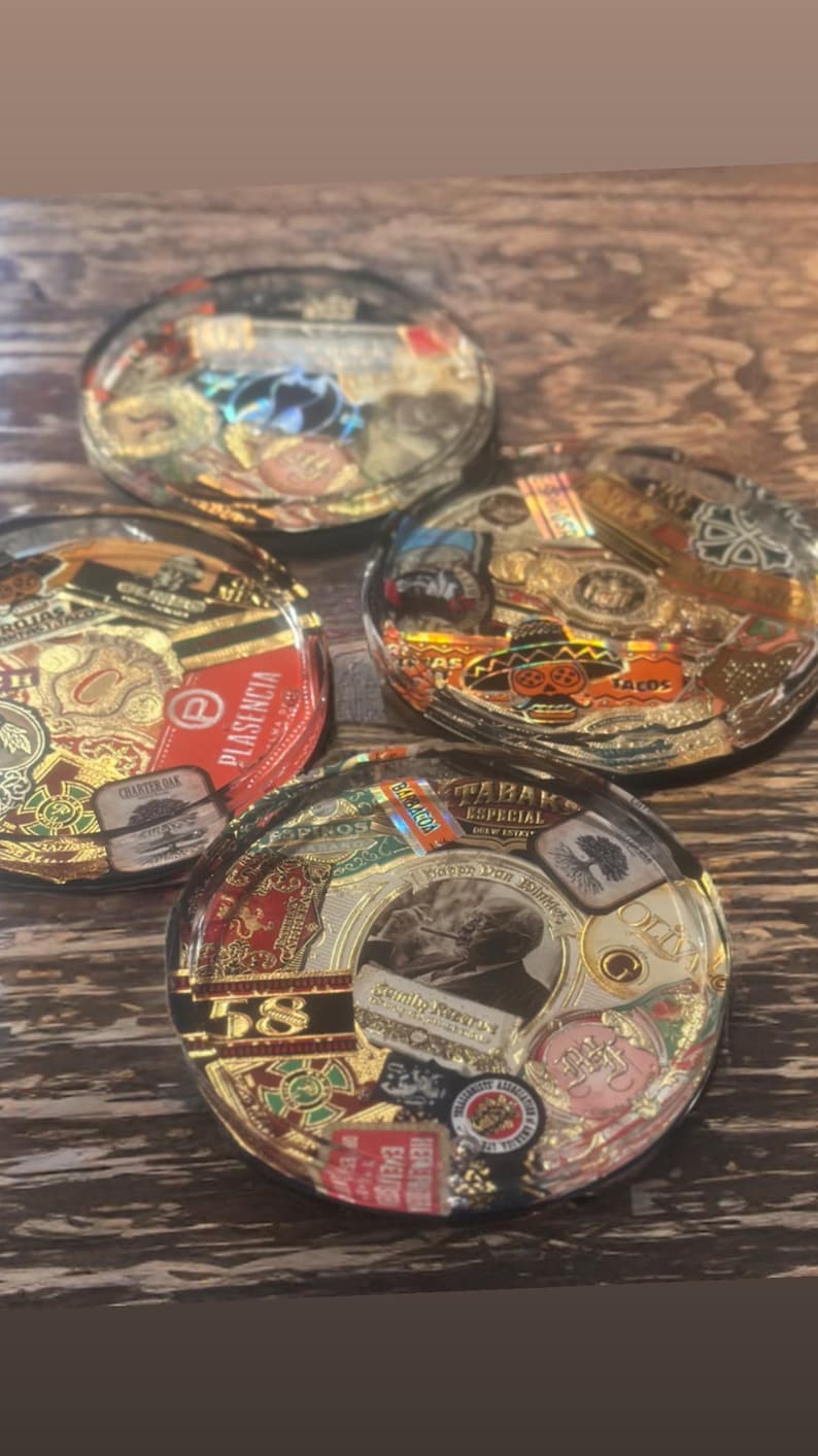 Puede incluir: Cuatro posavasos de resina transparente con dise&ntilde;os de etiquetas de cigarros. Cada posavasos presenta una etiqueta diferente con detalles dorados, rojos y marrones. Los posavasos est&aacute;n dispuestos sobre una superficie de madera.