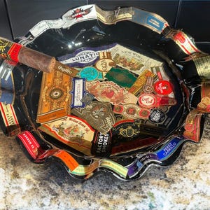 K&ouml;nnte beinhalten: Ein runder, schwarzer Aschenbecher aus Glas mit gewelltem Rand, verziert mit zahlreichen bunten Zigarrenringen, die im Glas eingebettet sind. Eine einzelne braune Zigarre liegt auf dem Rand. Die Ringe zeigen verschiedene Markennamen und Designs.