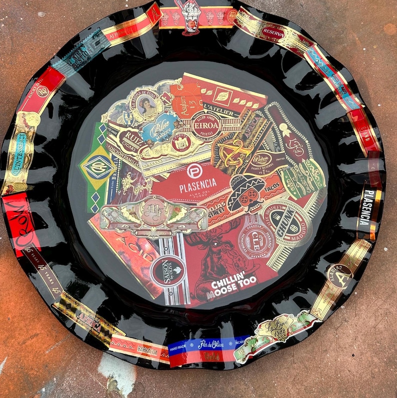 K&ouml;nnte beinhalten: Ein runder Aschenbecher aus schwarzem Glas mit einer Collage aus verschiedenen bunten Zigarrenringen, die unter der Oberfl&auml;che eingebettet sind. Die Mitte zeigt Etiketten wie Plasencia und Chillin' Moose Too, w&auml;hrend der gewellte Rand mit weiteren Logos verziert ist.