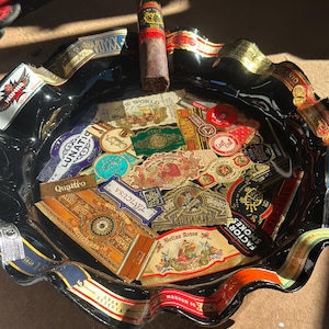 K&ouml;nnte beinhalten: Ein schwarzer Aschenbecher aus Glas mit gewelltem Rand, der eine Collage aus verschiedenen bunten Zigarrenringen auf der Oberfl&auml;che zeigt. Eine einzelne braune Zigarre mit einem rot-goldenen Ring liegt auf dem oberen Rand des Aschenbechers, der zahlreiche Markennamen und komplexe grafische Designs aufweist.